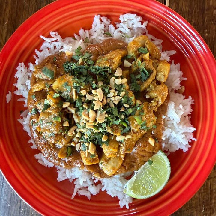 Satay chicken on jasmine rice with chopped peanuts, coriander and a lime wedge, on a red plate.